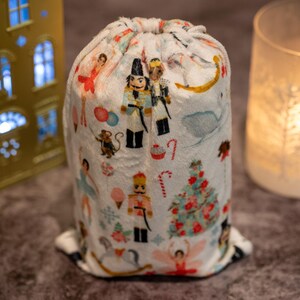 Puede incluir: Una bolsa de cordón blanca con un estampado navideño con cascanueces, bailarinas, bastones de caramelo y un árbol de Navidad. La bolsa está hecha de una tela suave y de felpa.
