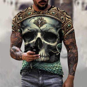 Puede incluir: Una camiseta de manga corta negra y verde con un estampado gráfico de una calavera con una serpiente enroscada a su alrededor. La calavera está rodeada de un marco dorado con gemas verdes.