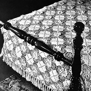 May include: A black and white image of a bed with a detailed, white crocheted bedspread. The bedspread has a geometric pattern and a fringed edge. The dark wooden bed frame is visible.