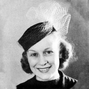 May include: A black and white photo of a woman wearing a black hat with a white veil. The hat is tilted on her head and the veil is draped over her hair. She is smiling and looking at the camera.