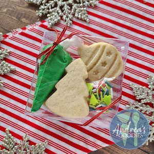 May include: A clear plastic box tied with a red ribbon contains Christmas-themed cookies and decorating supplies. The cookies are shaped like a Christmas tree and an ornament. A green icing bag and colorful sprinkles are also included.