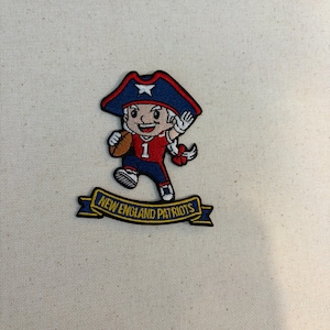 May include: An embroidered patch featuring a cartoon character wearing a blue tricorn hat with a white star, a red jersey with the number 1, and blue pants. The character holds a brown football. Below is a blue banner with New England Patriots text.
