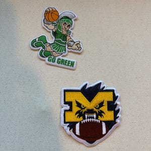 May include: Two embroidered patches. The top patch features a green and white Spartan mascot holding an orange basketball with the text GO GREEN. The bottom patch displays a yellow letter M with a black creature face and a brown football.