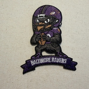 May include: An embroidered patch featuring a cartoon raven mascot wearing a purple football helmet and jersey, holding a brown football. Below the character is a purple banner with the white text BALTIMORE RAVENS stitched in capital letters.