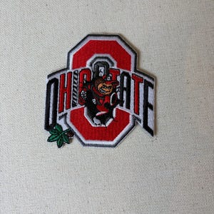 May include: An embroidered patch featuring a red block letter O with a white and black border. The text OHIO STATE is written in black across the center. A small brown and black mascot figure is centered, and a green leaf icon is at the bottom left.