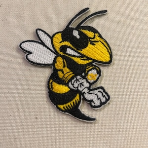 May include: Embroidered patch featuring a stylized yellow and black hornet with white wings. The hornet is depicted in a dynamic pose, with its arms clenched. The patch is designed with detailed stitching and a clean outline, suitable for application on various fabrics.