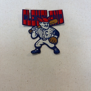 May include: An embroidered patch featuring a cartoon baseball player in a white pinstripe uniform with a blue C on the chest, wearing a red cap with wings, holding a brown bat, and a red banner above with the blue text CLEVELAND.