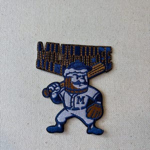 May include: An embroidered patch featuring a cartoon baseball player character with a beer mug head, wearing a white uniform with a blue M, holding a wooden baseball bat, with the word MILWAUKEE in blue block letters positioned above the character.