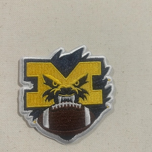 May include: Embroidered patch featuring a yellow letter "M" with a snarling wolverine face, a navy blue outline, and a brown football at the bottom. The patch has a white border.