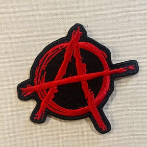 May include: Embroidered patch featuring the anarchy symbol. The design has a red "A" inside a circle, with red lines extending outward, all on a black background. The patch has a black border.