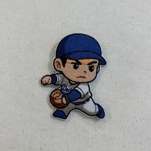 May include: Embroidered patch featuring a cartoon baseball player in a blue cap and uniform, holding a baseball. The patch has a white background and is likely for apparel or accessories.