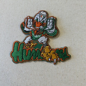 May include: Embroidered patch featuring a stylized mascot in green, orange, and white, with the word "Hurricanes" in green and orange. The patch has a detailed design and is likely for apparel.