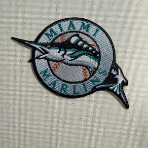May include: Embroidered patch featuring the Miami Marlins logo. The patch has a circular design with the words "MIAMI MARLINS" in teal lettering. The logo includes a teal marlin leaping over a baseball, with orange and black accents.