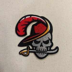 May include: Embroidered patch featuring a skull wearing a pirate hat with a red feather and a sword in its mouth. The skull is gray with black outlines and details, and the hat has yellow and black accents.
