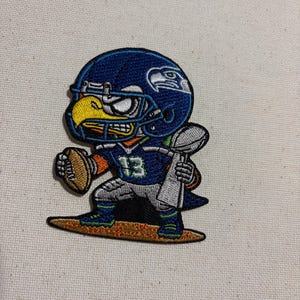 May include: Embroidered patch featuring a cartoon bird in a football uniform. The bird wears a blue helmet with a white logo and holds a football and a trophy. The number 13 is visible on the jersey.