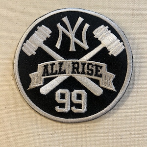 May include: A circular black and white patch featuring the New York Yankees logo, crossed hammers, and the text "ALL RISE 99". The patch has a white border and is likely an embroidered item.