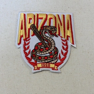 May include: An embroidered patch featuring the word ARIZONA in large yellow letters with red outlines. Below the text is a coiled rattlesnake with a brown and tan pattern, positioned next to a brown stick. A red laurel wreath and the year 1998 are below.