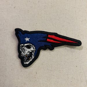 May include: Embroidered patch featuring a skull design within a blue and red stylized shape. The patch has a black border and a white star. The design is reminiscent of a team logo.