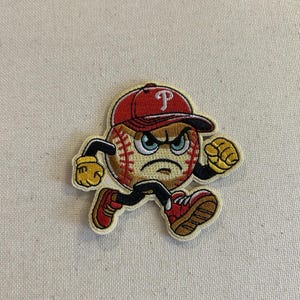 May include: Embroidered patch featuring a baseball with an angry face, wearing a red baseball cap with a white "P". The baseball has arms and legs, wearing red shoes and yellow gloves, in a running pose. The patch is on a beige background.