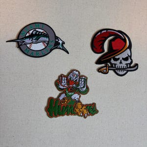 May include: Three embroidered patches featuring sports team logos. One is a circular Miami Marlins logo with a marlin and baseball. Another is a skull wearing a red feathered hat with a sword. The third is a duck mascot with the text Hurricanes.