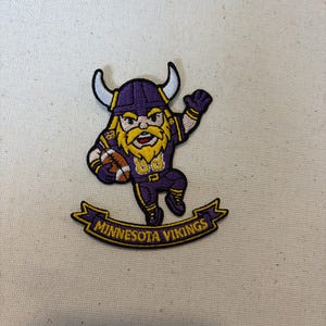 May include: An embroidered patch featuring a cartoon Viking character wearing a purple helmet with white horns, a purple jersey with yellow accents, and holding a football. Below the character is a purple banner with the text MINNESOTA VIKINGS.