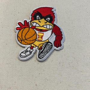 May include: Embroidered patch featuring a cartoon cardinal bird in a basketball uniform, holding a basketball. The bird is red, yellow, and white, with black accents. The patch has a white border and is on a beige background.
