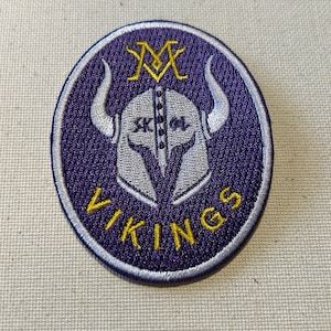May include: A circular purple patch with a white and yellow Viking helmet design. The patch has the word "VIKINGS" in yellow text at the bottom and the letter "M" above the helmet. The word "SKOL" is visible on the helmet.