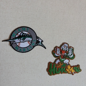 May include: Two embroidered patches. The left patch is a circular Miami Marlins logo featuring a marlin fish over a baseball. The right patch depicts a cartoon duck character in orange, green, and white, positioned above the green text "HURRICANES".