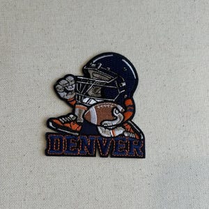 May include: Embroidered patch featuring a football player in a navy blue helmet, holding a football. The patch has orange and white accents, with the word "DENVER" in navy blue letters at the bottom. The patch is on a beige background.