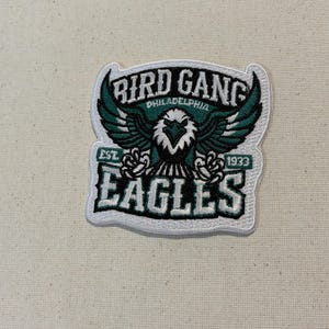 May include: An embroidered patch featuring a central eagle with spread wings in dark green, white, and black. The text reads BIRD GANG, PHILADELPHIA, EST. 1933, and EAGLES. The design is outlined in white thread on a white fabric background.