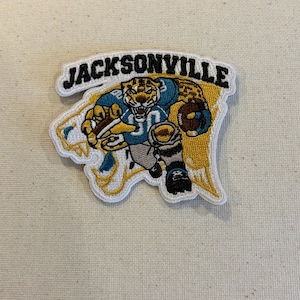 May include: An embroidered patch featuring a cartoon jaguar mascot in a teal and gold football uniform holding a brown football. The word JACKSONVILLE is stitched in black capital letters across the top of the patch, which has a white border.