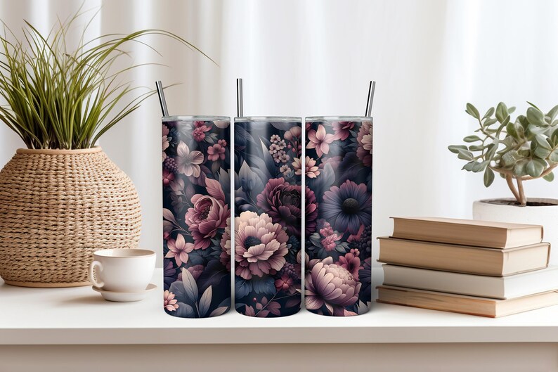 Puede incluir: Tres vasos de acero inoxidable con dise&ntilde;os florales. Los vasos presentan un fondo oscuro con flores rosas, moradas y blancas. Cada vaso tiene una pajita plateada. Los vasos se exhiben sobre una superficie blanca.