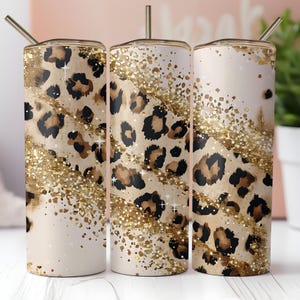 May include: A set of three stainless steel tumblers with lids and straws. The tumblers feature a beige background with a leopard print design in brown and black. Gold glitter accents and a gold rim complete the design.