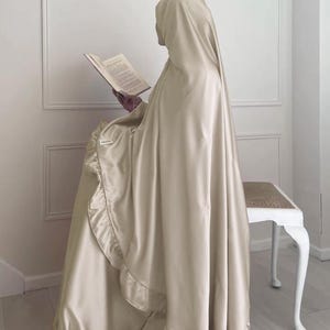 May include: A beige satin prayer shawl with a ruffled trim. The shawl is draped over a chair and a book is resting on the chair.