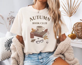 May include: Cream-colored t-shirt with the text "AUTUMN BOOK CLUB" in brown. The graphic features a coffee mug, book, candle, leaves, and glasses. The shirt is paired with denim shorts and a beige cardigan.