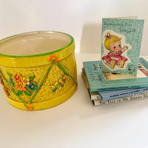 May include: A yellow ceramic planter with a textured surface and raised floral decorations, accompanied by a stack of three children's books and a greeting card that reads Announcing Our Baby Girl with an illustration of a baby.