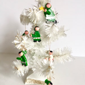 Op de afbeelding: Een kleine witte kerstboom versierd met houten ornamenten. De ornamenten tonen figuren in groene en witte kleding, zwarte schoenen en roze wangen. Een gouden ster en een gouden bal bekronen de boom.