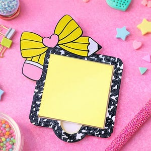 May include: A novelty notepad shaped like a composition notebook with a yellow pencil and pink eraser design. The notepad has a yellow sticky note pad and a black and white border. The pencil has a pink heart in the centre of the bow.