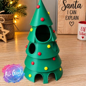 May include: A green, cone-shaped Christmas tree ornament featuring multiple circular openings and small, round red and yellow decorative accents. The structure has a tiered design with arched cutouts at the base, resembling a stylised tree.
