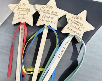May include: Wooden star-shaped graduation wands with personalized text. Each wand features a star with school names and "Graduate" text, plus the year "2025". Ribbons in red, blue, yellow, green, and white are attached. The wands are for Woodburn Pre-School, Sunnylands Nursery School, and Oakfield Nursery Unit.