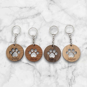 May include: Four wooden keyrings with paw print cutouts and personalised names: Daisy, Oscar, Remy, and Jasper. The keyrings are brown and have silver key rings.