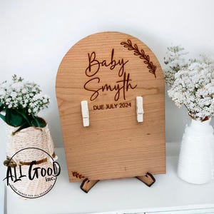 May include: A wooden sign with a rounded top and a stand. The sign is engraved with the text "Baby Smyth" and "Due July 2024".