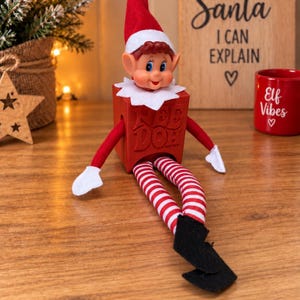 May include: A toy elf with a red hat, white collar, and red and white striped legs, sitting inside a red square container embossed with the text 'PEE DOH'. The elf has white mittens and black pointed shoes, with a smiling face and pointed ears.
