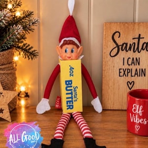 May include: A plush elf doll wearing a red hat with a white tip, red long sleeves, white mittens, and red and white striped leggings with black shoes. The elf holds a yellow rectangular box labelled .4oz Santa's BUTTER in blue text.