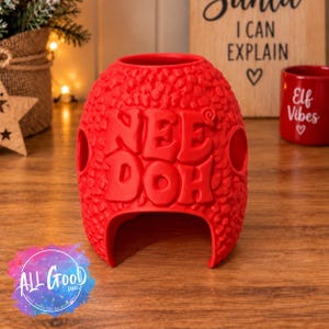 May include: A bright red, textured silicone Nee Doh stress ball cover featuring a bumpy surface and the embossed text Nee Doh on the front. The object has a hollow, rounded shape with a large arched opening at the bottom and two side holes.
