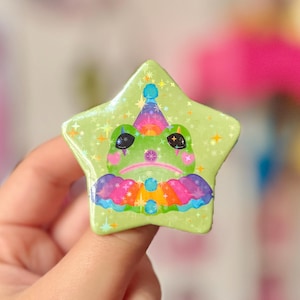 May include: A star-shaped pin featuring a sad frog face wearing a rainbow party hat and collar. The pin is light green with glitter and star accents. The frog has black eyes and a pink mouth. The pin is held by a hand with pink nail polish.