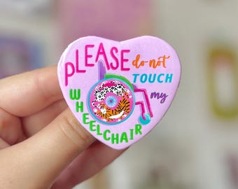 May include: A heart-shaped button with the text "PLEASE do not TOUCH my WHEELCHAIR" in pink, orange, green, and blue. The button features a colorful wheelchair design with a tiger print wheel.