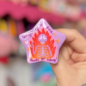 May include: A star-shaped badge with a purple background and a skeleton design in flames. The text "everything is fine, thanks for asking!" is written on the badge.