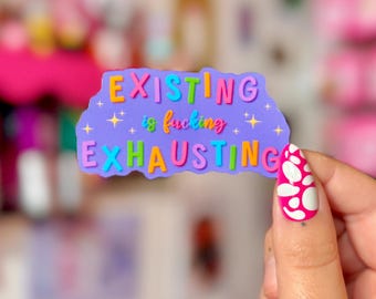 May include: A purple sticker with the text "EXISTING is fucking EXHAUSTING" in colorful bubble letters. The sticker has a wavy edge and small star accents. The image also shows a hand with a pink nail with white polka dots.