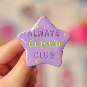 May include: A lavender star-shaped badge with the text "ALWAYS in pain CLUB" in pink, green, and blue. The badge has yellow star accents. The badge is held by a person.